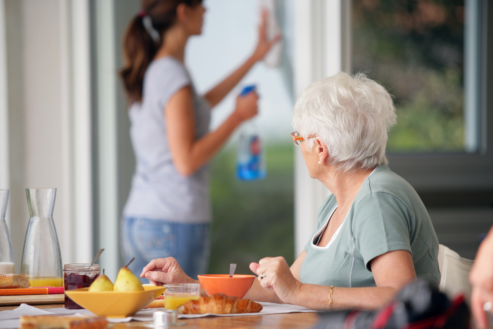 an elderly woman eating breakfast while a home care worker cleans in the background
